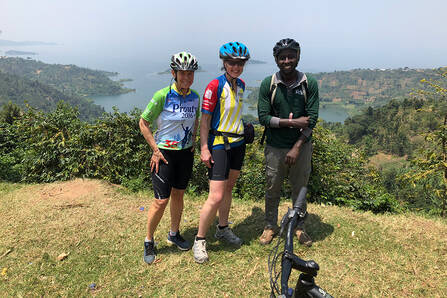 Three people dressed in bicycling clothes and helmets, standing on a grassy rise over a harbor view in Rwanda